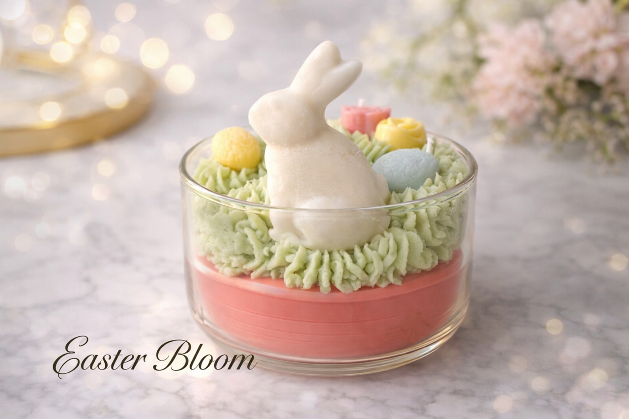 Easter Artisan Themed soy wax candle. Easter Bloom written on image. Clear glass vessel with pink base, green wax on top resembling grass, ivory wax easter bunny, adorned with decorative wax floral and egg melts. 