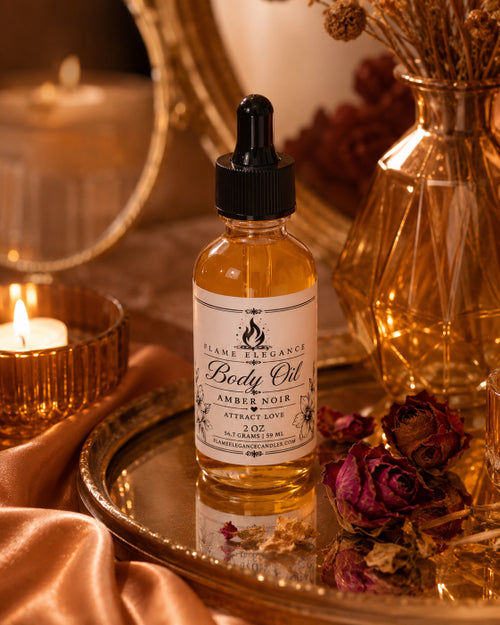 A bottle of Flame Fragrance Body Oil in the Ambre Noir scent, styled on a gold tray with dried roses, a lit candle, and warm ambient lighting.