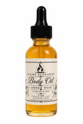 A 2 oz amber glass dropper bottle of Flame Elegance Amber Noir Body Oil in the Amber Noir scent, featuring a cream-colored label with black text and a black dropper cap, set against a plain white background.