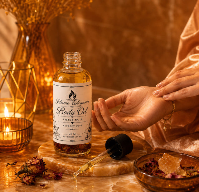 A bottle of Flame Elegance Body Oil with a dropper lies on a marble slab in a warm, candlelit setting, with a person's hands reaching toward it and decorative dried flowers and a bowl nearby.