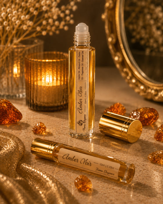 Two amber roll-on perfume bottles with gold caps and cream labels reading 'Amber Noir' and 'Tome Elegance', styled on a warm stone surface with a lit candle, dried flowers, gold fabric, and amber crystals by Flame Elegance