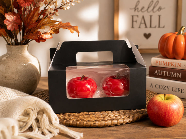 Two scented apple bliss candles in black packaging by Flame Elegance sitting on brown hot pad with fall flowers in background and a apple and pumpkin