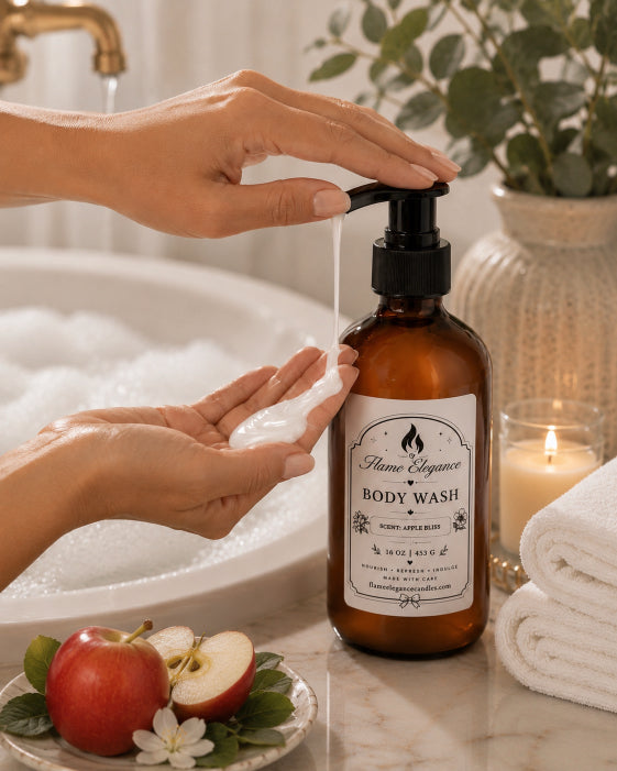 An amber plastic body wash bottle with a black pump and a white label reading 'Flame Elegance Apple Bliss BODY WASH' is being dispensed with hands in front of a bubble bath, with apples, a candle, and folded towels on a marble surface.
