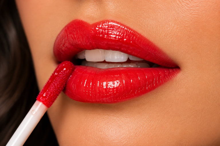 Close-up of glossy bright red lipstick being applied with a doe-foot applicator to a person's lips.