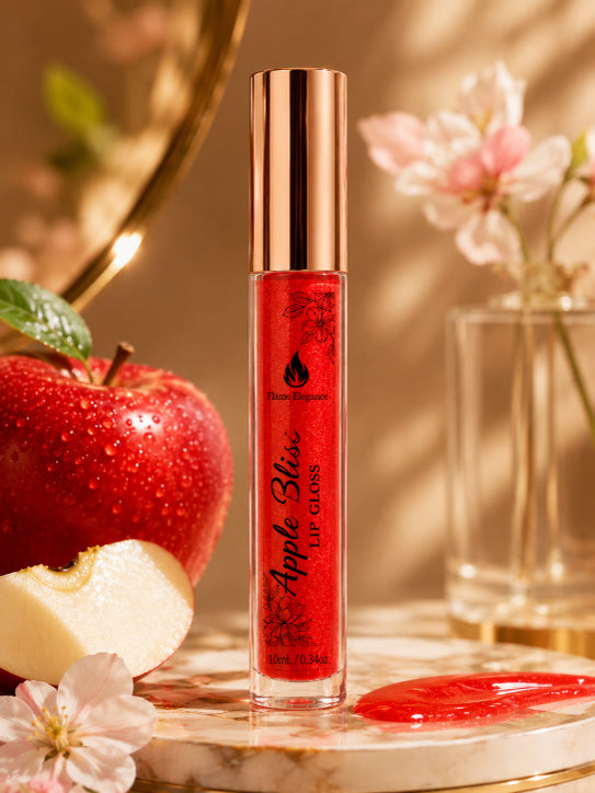 A tube of red apple-bliss lip gloss by Flame Elegance stands on a marble surface with a glossy red smear in front, surrounded by a red apple, a sliced apple, and soft pink flowers in a warm, sunlit setting.