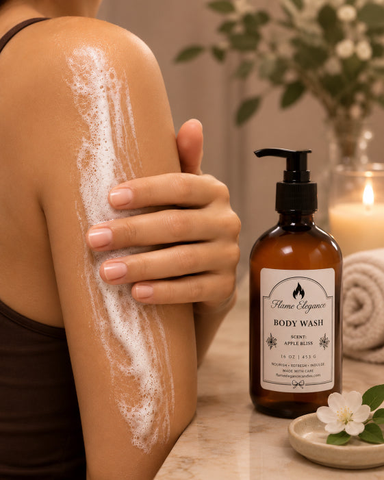 A person applies white Apple Bliss body wash soap by Flame Elegance to their upper arm while standing beside an amber pump bottle labeled Home Vignette Body Wash on a marble surface, with a lit candle and greenery in the background.