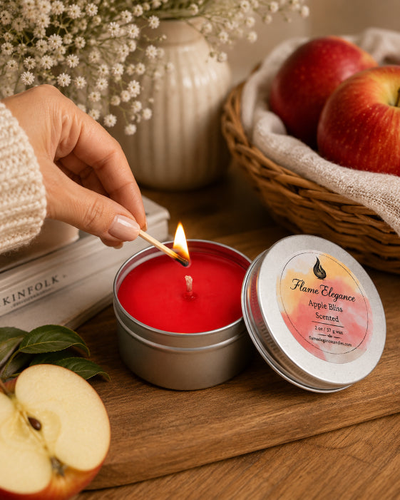 A red scented candle in a silver tin is lit with a wooden match by a person's hand. The silver lid sits beside the candle on a wooden table, with an apple half, a wicker basket of red apples, and a cream ribbed vase in the background.