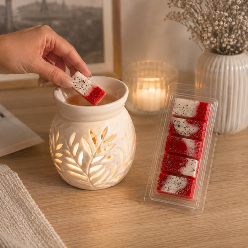 Hand dipping a Apple Bliss elegant wax melt into a scented warmer on a wooden surface with packaging and candles in the background by Flame Elegance.
