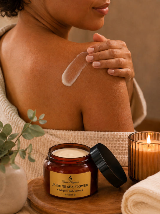 Person applying Luxe Jasmine Sea Flower Whipped Body Butter by Flame Elegance to shoulder with a jar of Jasmine Sea Flower body butter and lit candle in the background.