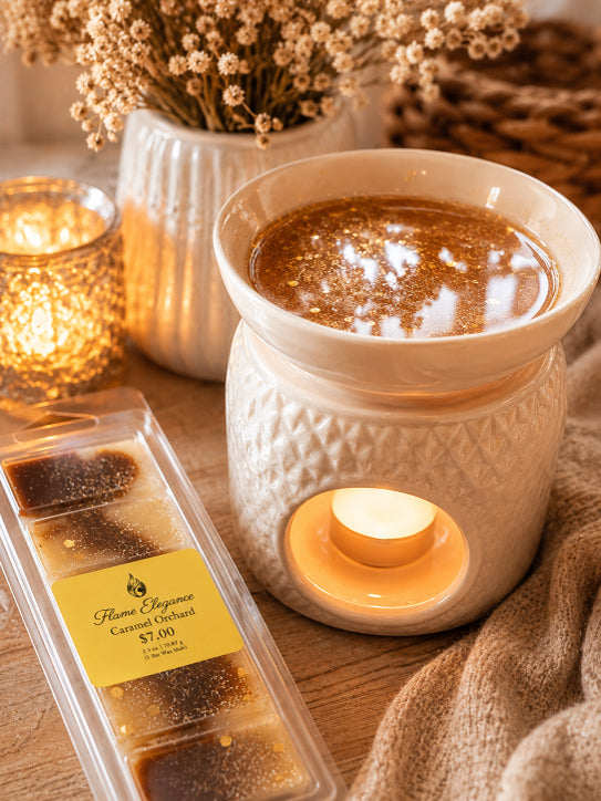 package of caramel orchard luxury soy wax melt bar with wax warmer in background lit with candle and shimmering caramel orchard wax melts melted in warmer by flame elegance. 