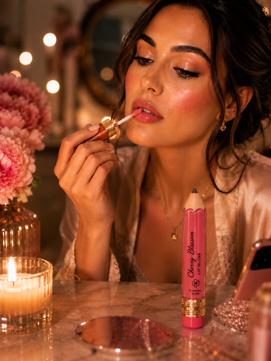 A woman applies pink lip gloss while a pink lip pencil labeled 'Cherry Blossom Lip Color' sits on a marble table beside a lit candle and flowers in a warm, romantic setting.