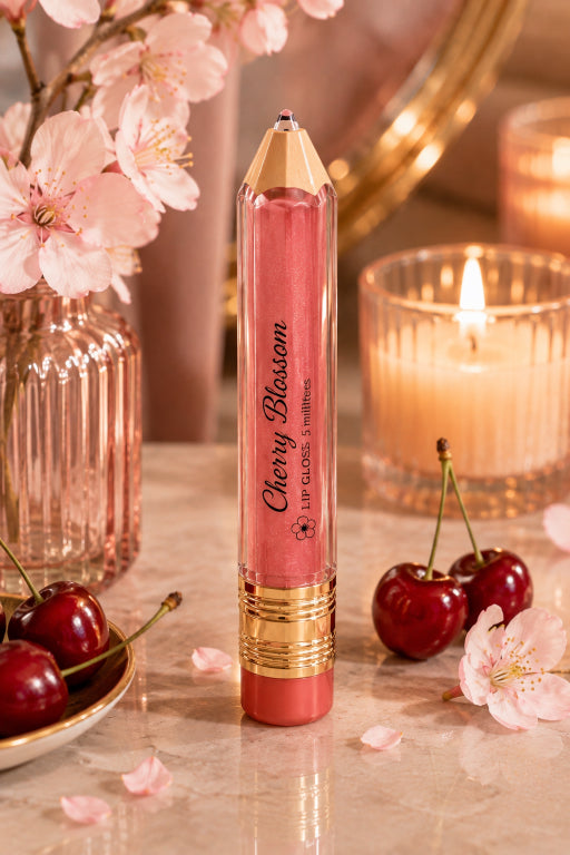 A lip gloss stick in a pencil-shaped tube labeled 'Cherry Blossom' stands on a marble surface surrounded by pink cherry blossoms, cherries, and lit candles. The product is pink with gold detailing and a coral-red tip.