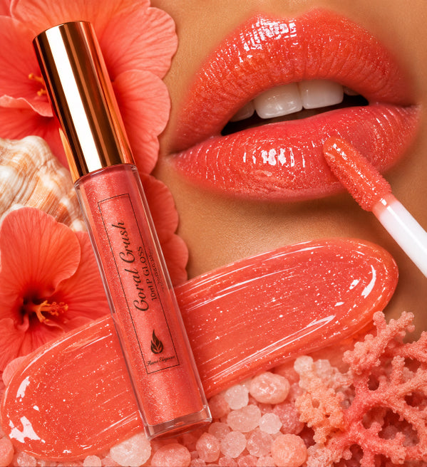 A close-up beauty shot showing a coral-pink lip gloss tube labeled 'Coral Crush Flame Elegance' alongside a doe-foot applicator on glossy lips, with coral-themed props and floral elements in the background.