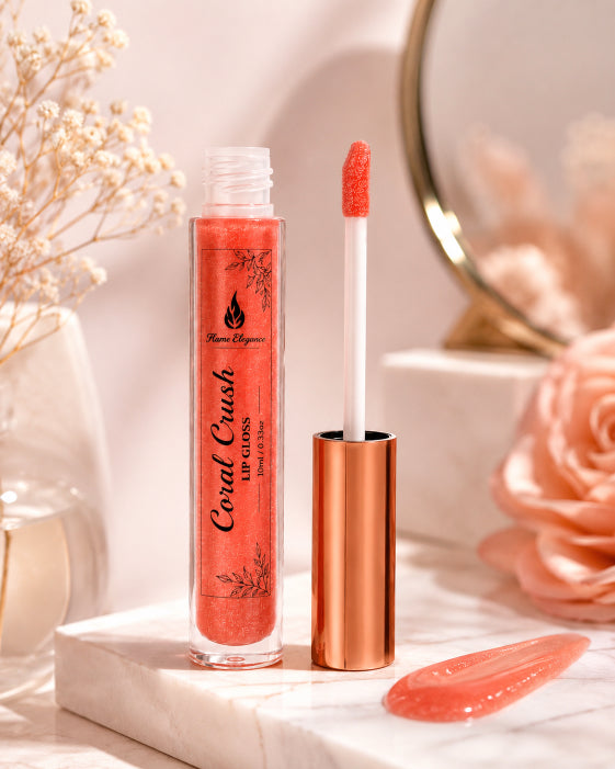 A tube of coral lip gloss with its cap off, standing next to the applicator wand on a white marble surface. The background is styled with dried flowers, a pink rose, and a mirror, creating a warm, soft aesthetic.