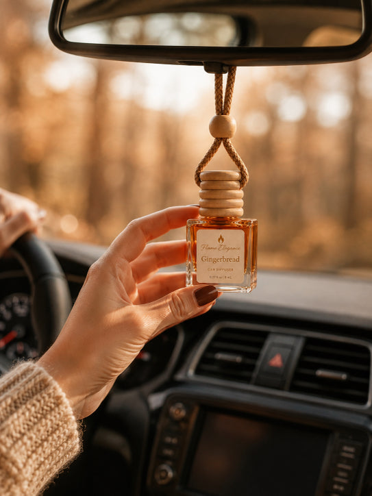 A hand holds a small glass car fragrance bottle labeled 'Gingerbread' with a wooden diffuser top, hanging from a rearview mirror inside a car with a warm, blurred outdoor background by Flame Elegance