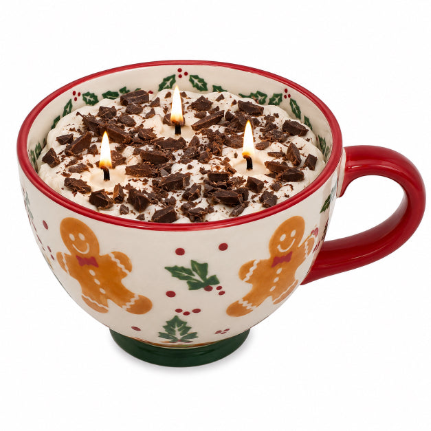 A ceramic mug-shaped candle with a white body decorated with gingerbread men, green holly leaves, and red dots, filled with white wax topped with chocolate shavings and three lit wicks, on a white background.