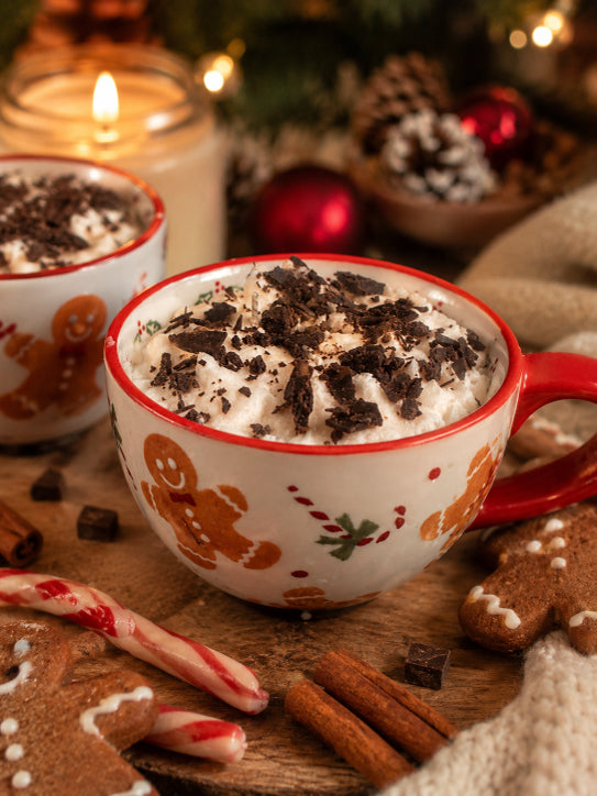 A festive holiday scene with two decorated ceramic mugs filled with whipped wax and chocolate wax crumbles, set on a wooden surface with gingerbread cookies, cinnamon sticks, candy canes, and a warm candle in the background. Soy wax candle by Flame Elegance.