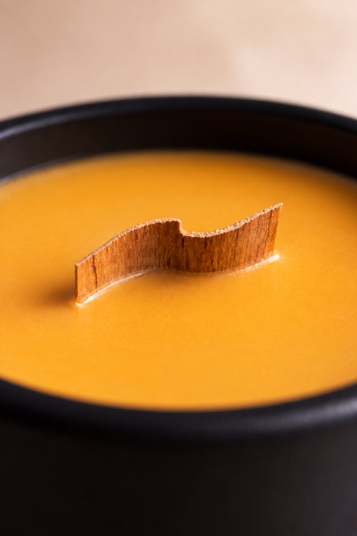 A close-up of a creamy orange-yellow soup or custard in a black bowl, with a small wooden stick or utensil standing upright in the center against a soft beige background.