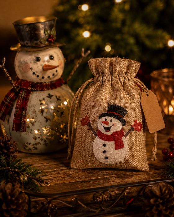 A small burlap drawstring gift bag with a printed snowman design stands on a wooden surface beside a decorative snowman figurine and a blurred Christmas tree in the background.