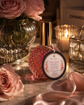A glittery pink bottle candle labeled 'Love Spell' by Flame Elegance styled on a marble surface with pink roses, a lit candle, and soft pink satin fabric.