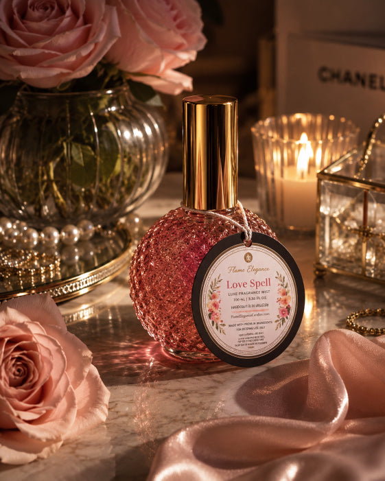 A glittery pink bottle candle labeled 'Love Spell' by Flame Elegance styled on a marble surface with pink roses, a lit candle, and soft pink satin fabric.