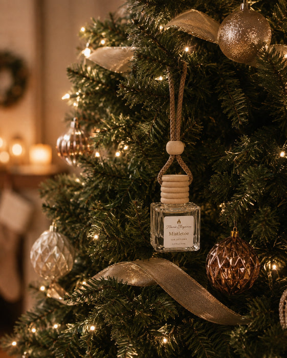 A Mistletoe scented Aroma Air Freshener diffuser hanging from a  Christmas tree  branches: a small square glass diffuser bottle with a reed diffuser-style top and a white label reading 'Mistletoe Flame Elegance. The tree is decorated with warm lights, gold ribbons, and metallic ornaments in a cozy indoor setting.