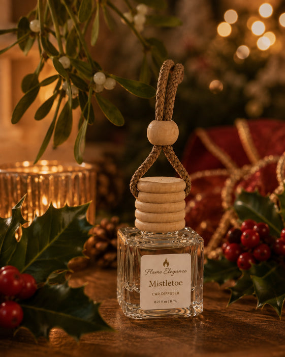 A small clear glass air freshener bottle labeled Flame Elegance Fragrance Mistletoe' sits on a wooden surface surrounded by holiday decor, including mistletoe, holly leaves and berries, and warm blurred lights in the background.