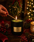 A black scented candle labeled 'Mistletoe' by Flame Elegance is lit with a wooden match in a cozy holiday setting. A hand with red nail polish holds the match near the flame.