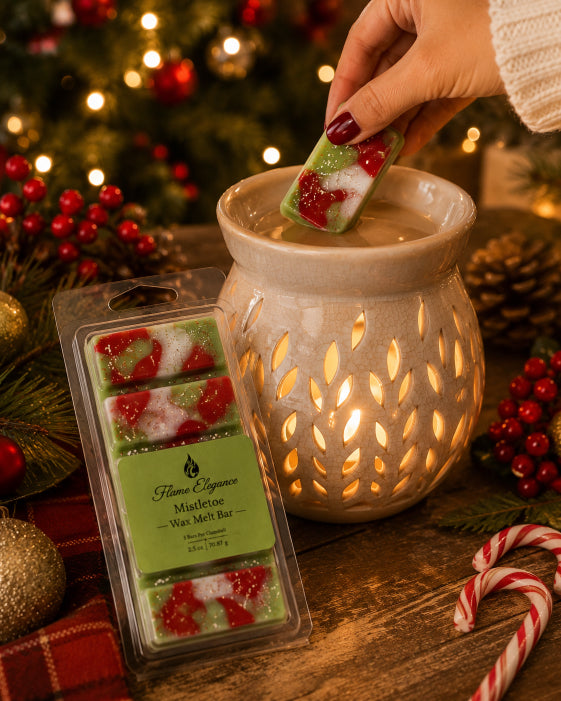 A hand holds a small wax melt bar above a lit ceramic wax warmer, with a package of holiday-themed wax melts on a wooden table surrounded by Christmas decorations including a pine tree, berries, and candy canes. Mistletoe soy wax melts by Flame Elegance in clamshell packaging.