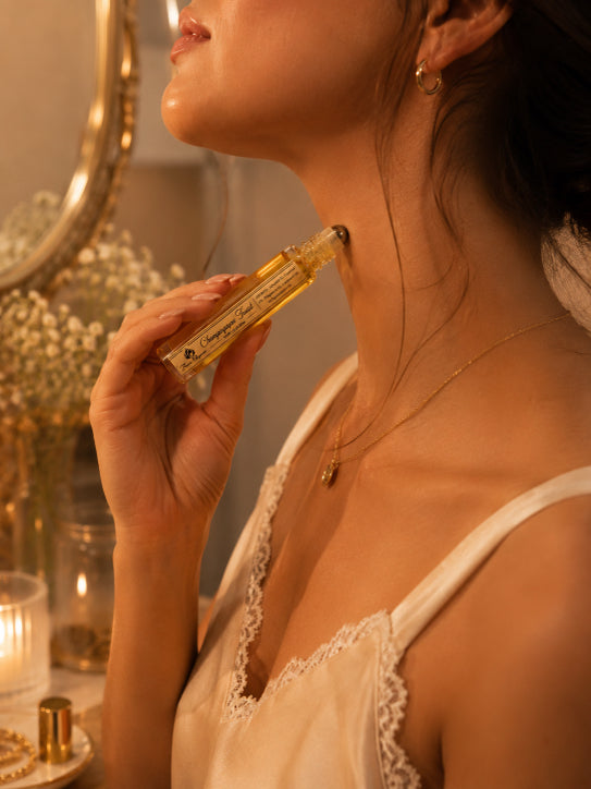 Woman applying Luxe Champagne Toast perfume with a mirror and decorative items in the background by Flame Elegance. 