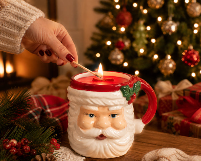 A hand lighting the Santa's hearth christmas wood wick candle that is housed in decorative Santa mug by Flame Elegance. Christmas tree lit in the background. Santa's Hearth Candle is sitting on a wood table. 