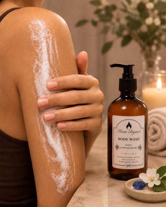 A person applies white Sapphire Desire body wash foam to their upper arm while standing beside an amber pump bottle labeled Home Superior Body Wash on a marble surface, with a lit candle, rolled towel, and flowers in the background by Flame Elegance.