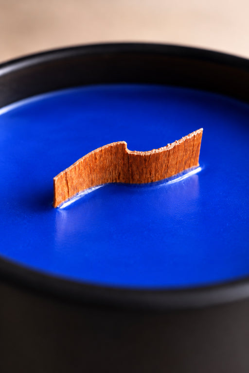 A close-up of a bright blue wax candle in a black container, with a decorative wavy wooden slice on the surface.