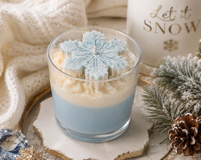 A layered candle in a clear glass jar sits on a decorative tray, surrounded by a cream knit blanket, a white candle with gold lettering reading 'Let it SNOW,' pine branches, and a pinecone. The candle has a light blue lower layer and a white upper layer with a light blue snowflake-shaped wax top. Snow kissed candle scented with sapphire desire by Flame Elegance