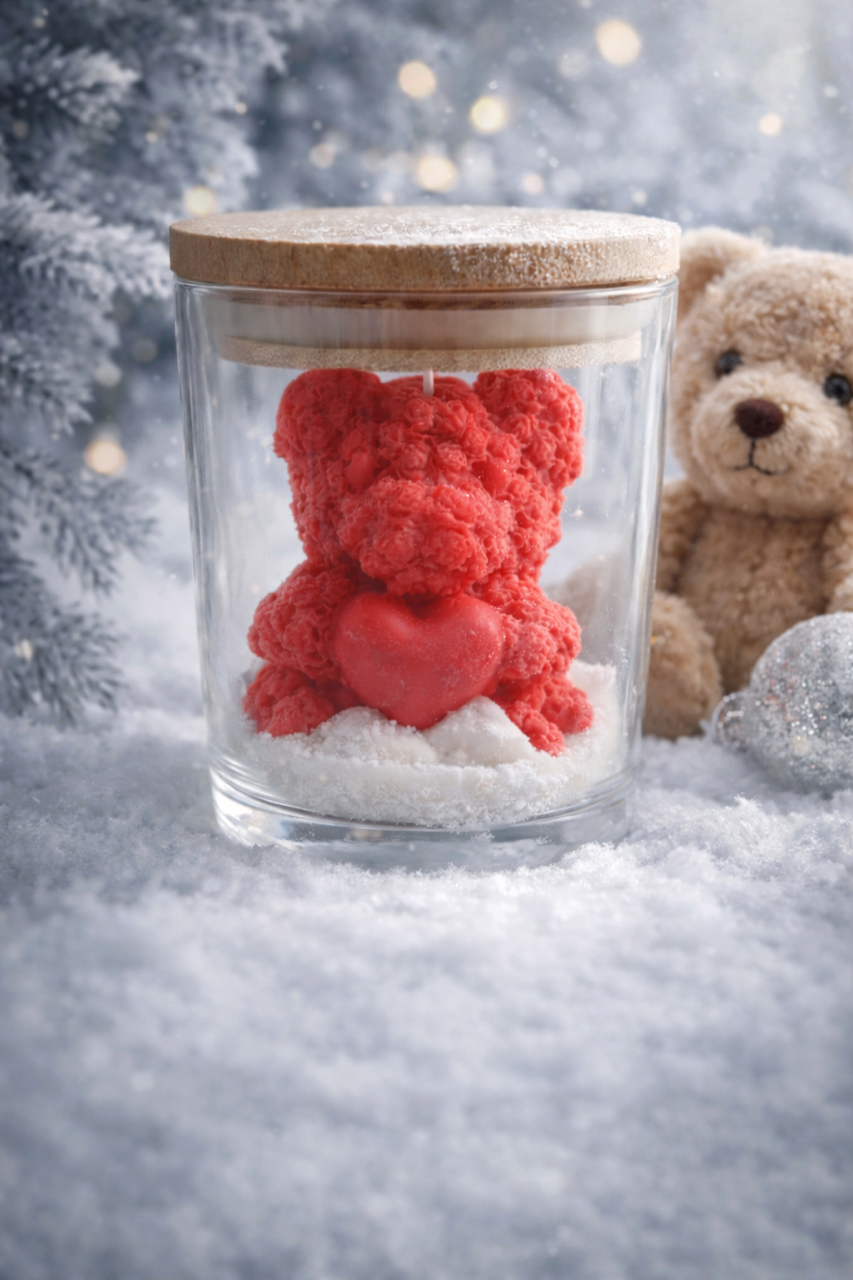 Red artisan candle teddy bear in a glass jar with a wooden lid, surrounded by soy wax that resembles snow and a blurred teddy bear in the background.