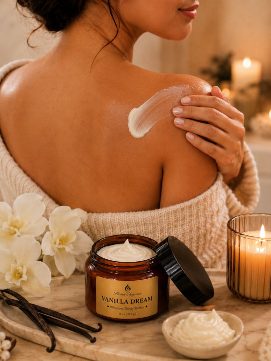 A woman in a cream knit sweater has a swipe of white body cream on her upper back. In the foreground, an open amber jar labeled 'Vanilla Dream' by The Spa Company sits on a wooden surface with vanilla beans, white flowers, and a lit candle by Flame Elegance.