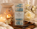 A packaged wax melt bar labeled Flame Elegance Vanilla Dream, shown standing upright on a wooden surface with candles, flowers, and vanilla beans around it.