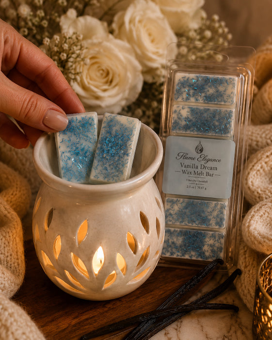A hand holds two blue wax melts with glitter above a white ceramic candle warmer, with a packaged box of Flame Elegance Vanilla Dream wax melts beside it on a wooden surface with flowers and a knit blanket in the background.