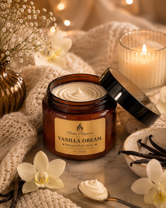An amber plastic jar of Flame Elegance Vanilla Dream whipped body butter with its black lid off, styled on a marble surface with a cream knit blanket, white orchids, vanilla beans, a spoon with cream, and a lit candle in the background.